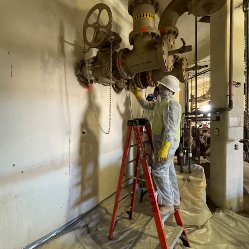 Worker in white protective suit inspecting industrial piping with valves in a utility room.