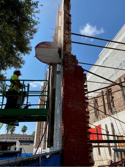 Worker on lift repairing a tall red brick wall beside a white building under a blue sky.