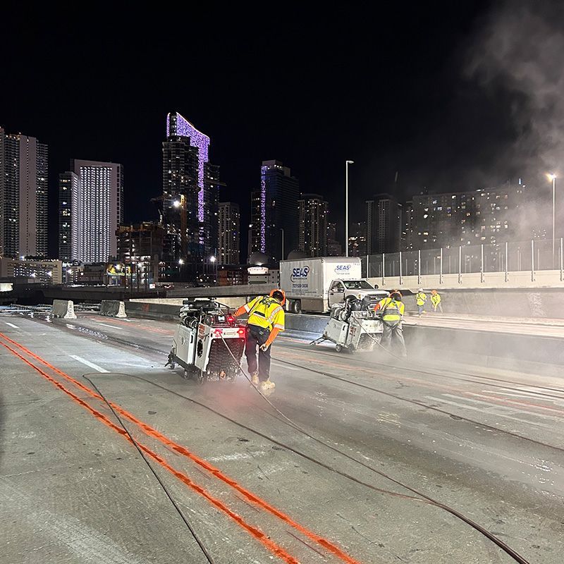 Road workers in reflective gear clearing snow on a city highway at night, with skyscrapers in the background