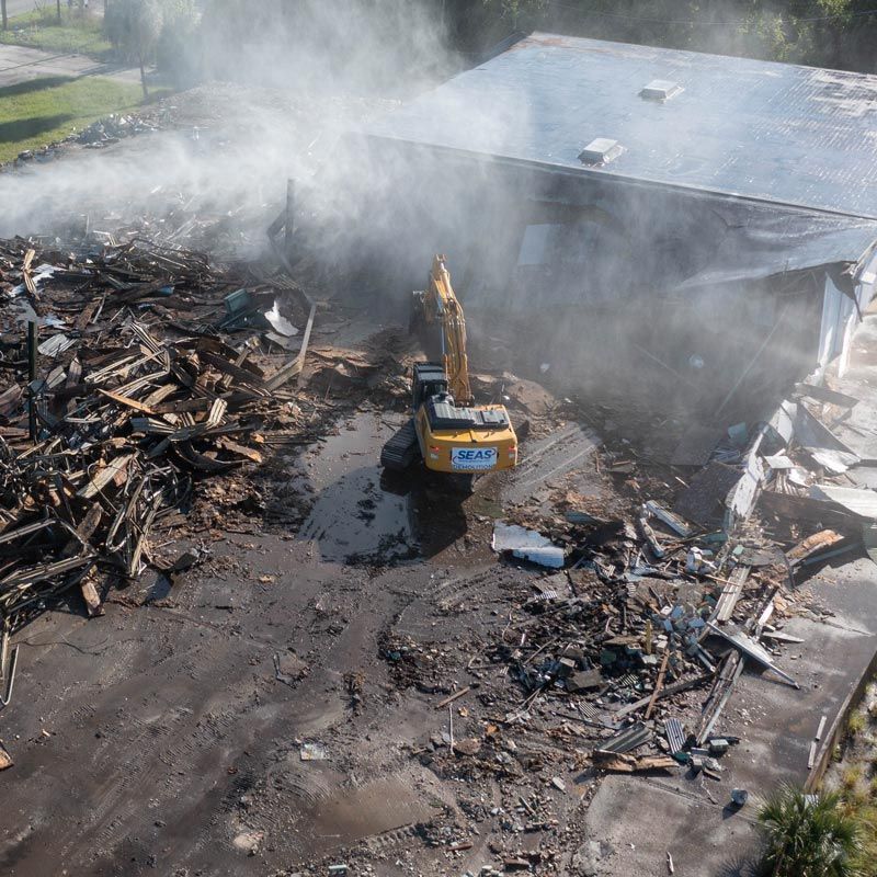 Excavator clearing debris at a smoky fire-damaged industrial site with a collapsed building.