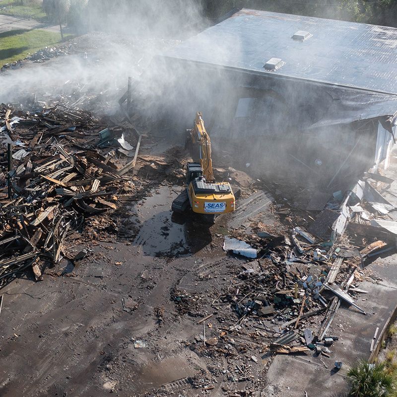 Excavator working amid smoking debris after a fire, beside a large metal building