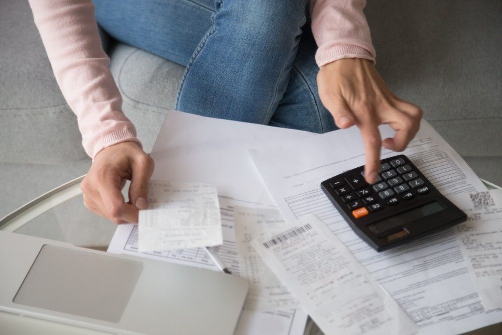 Person using a calculator surrounded by receipts and a laptop on a table, possibly budgeting.