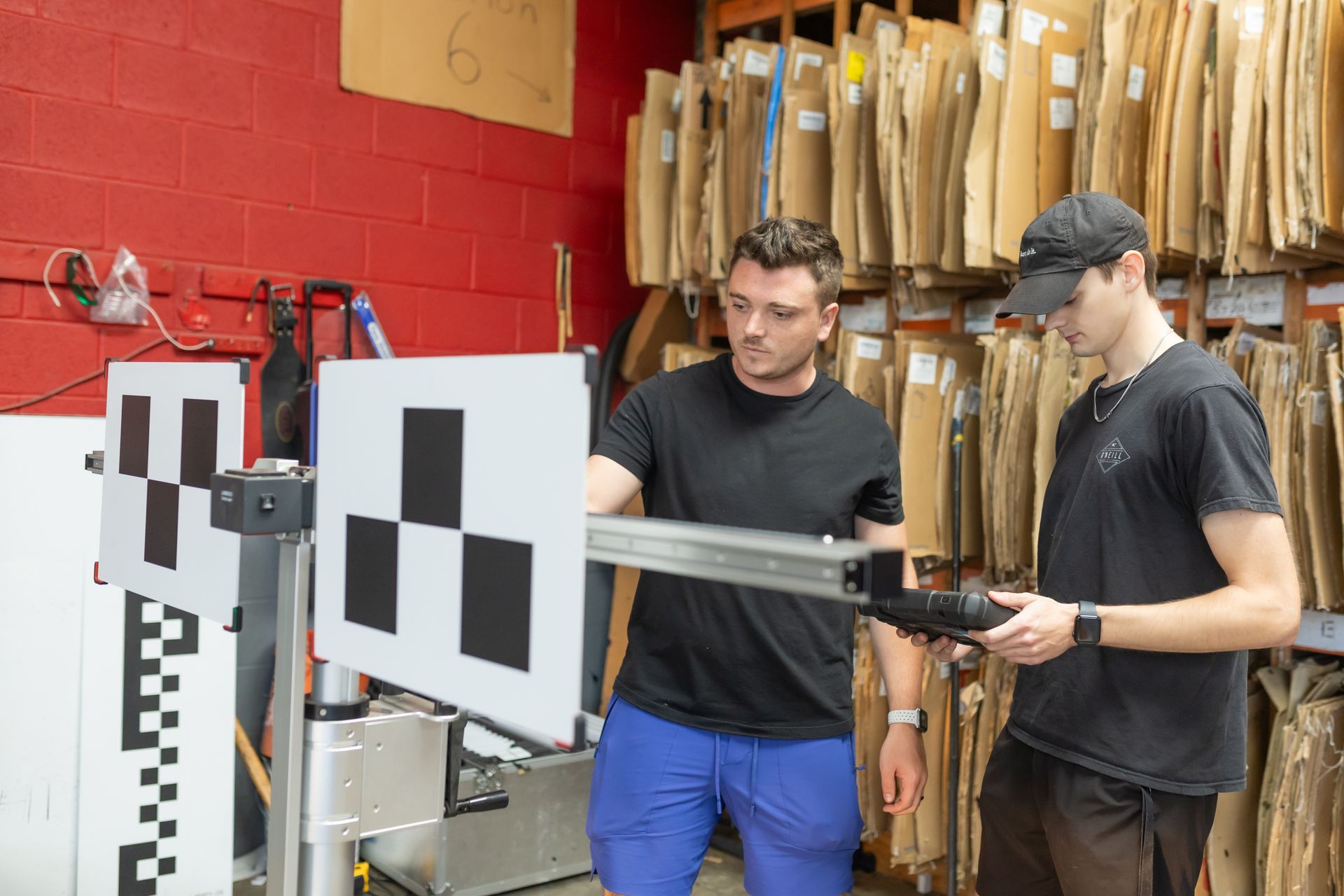 Two men in a workshop inspect a black and white grid target, using a camera and mounted equipment, likely for calibration.