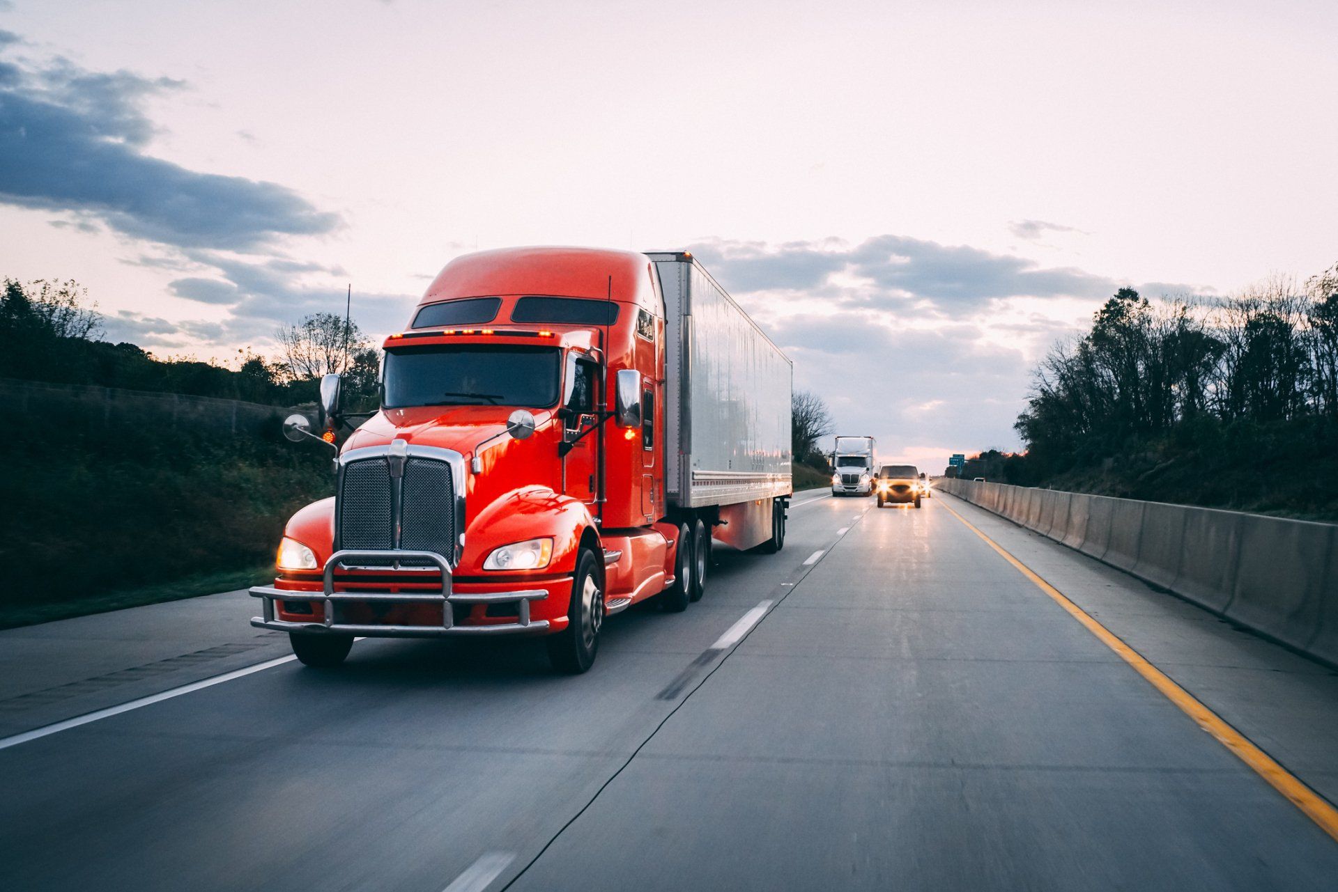 Red semi-truck driving on a highway at dusk. Other vehicles are visible in the distance.