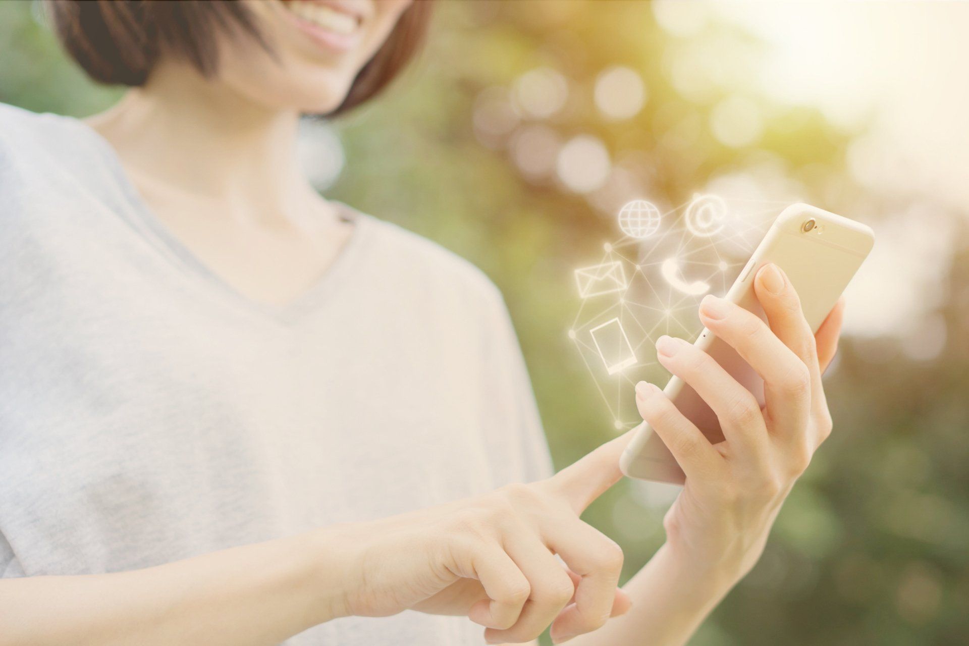 Woman smiling, using a gold smartphone, with digital communication icons above. Outdoor, blurred background.