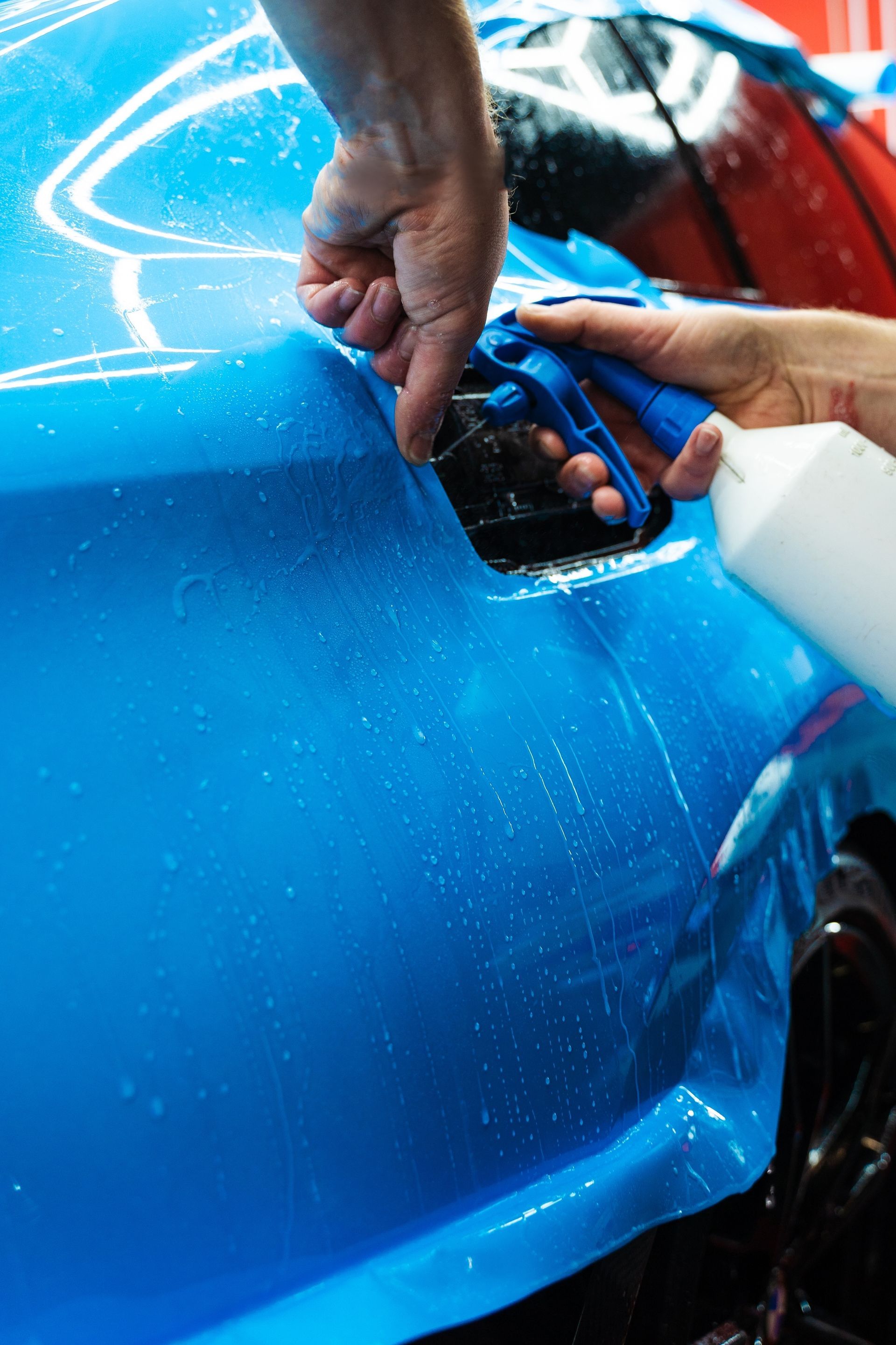 Hands peeling blue protective film from a car body panel at an auto shop