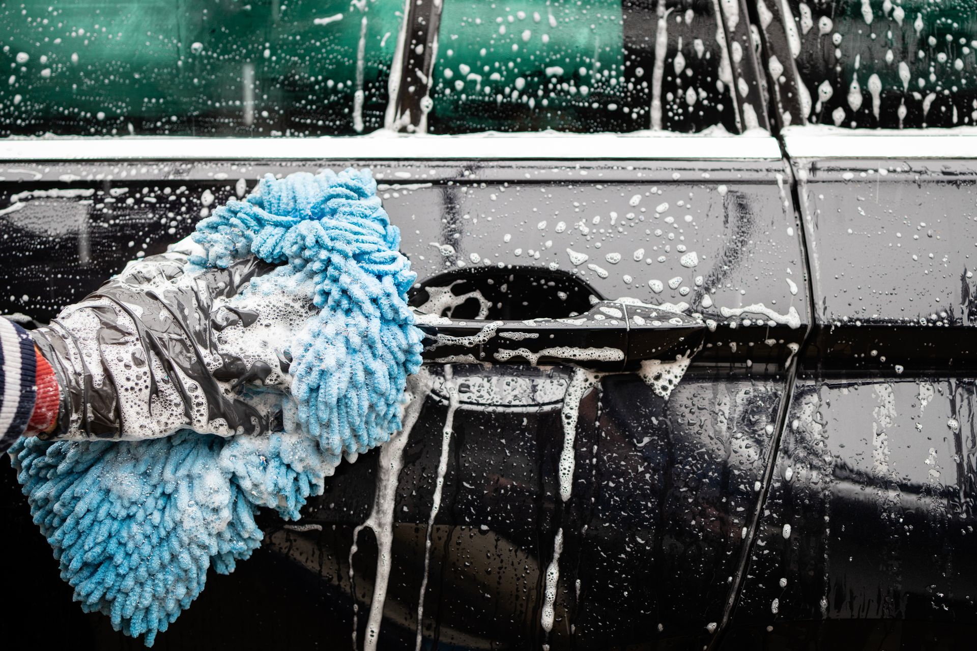 Gloved hand washing a black car door with blue sponge and soapy water splashing