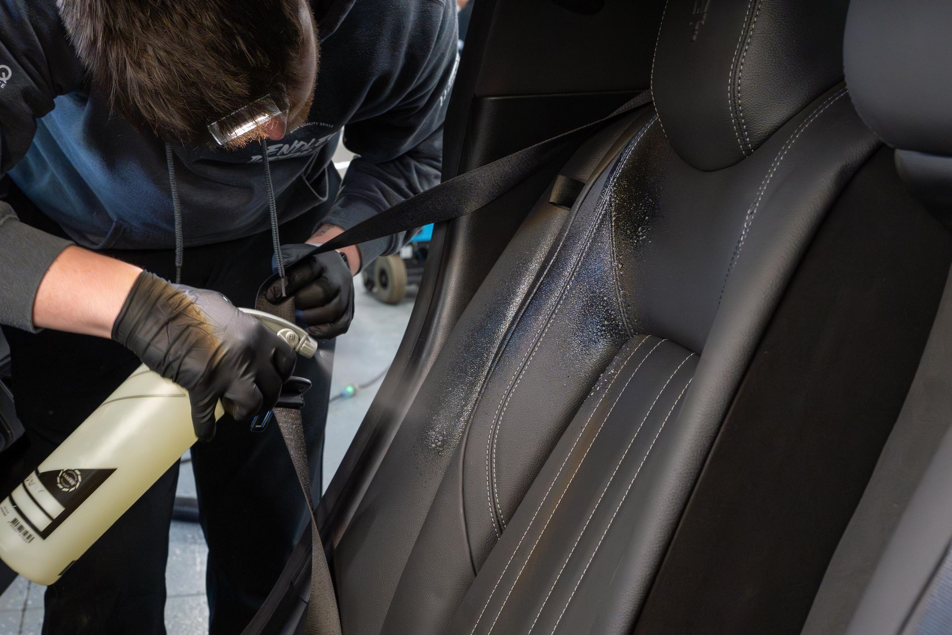Person cleaning a black car seat with a spray bottle and cloth inside a vehicle