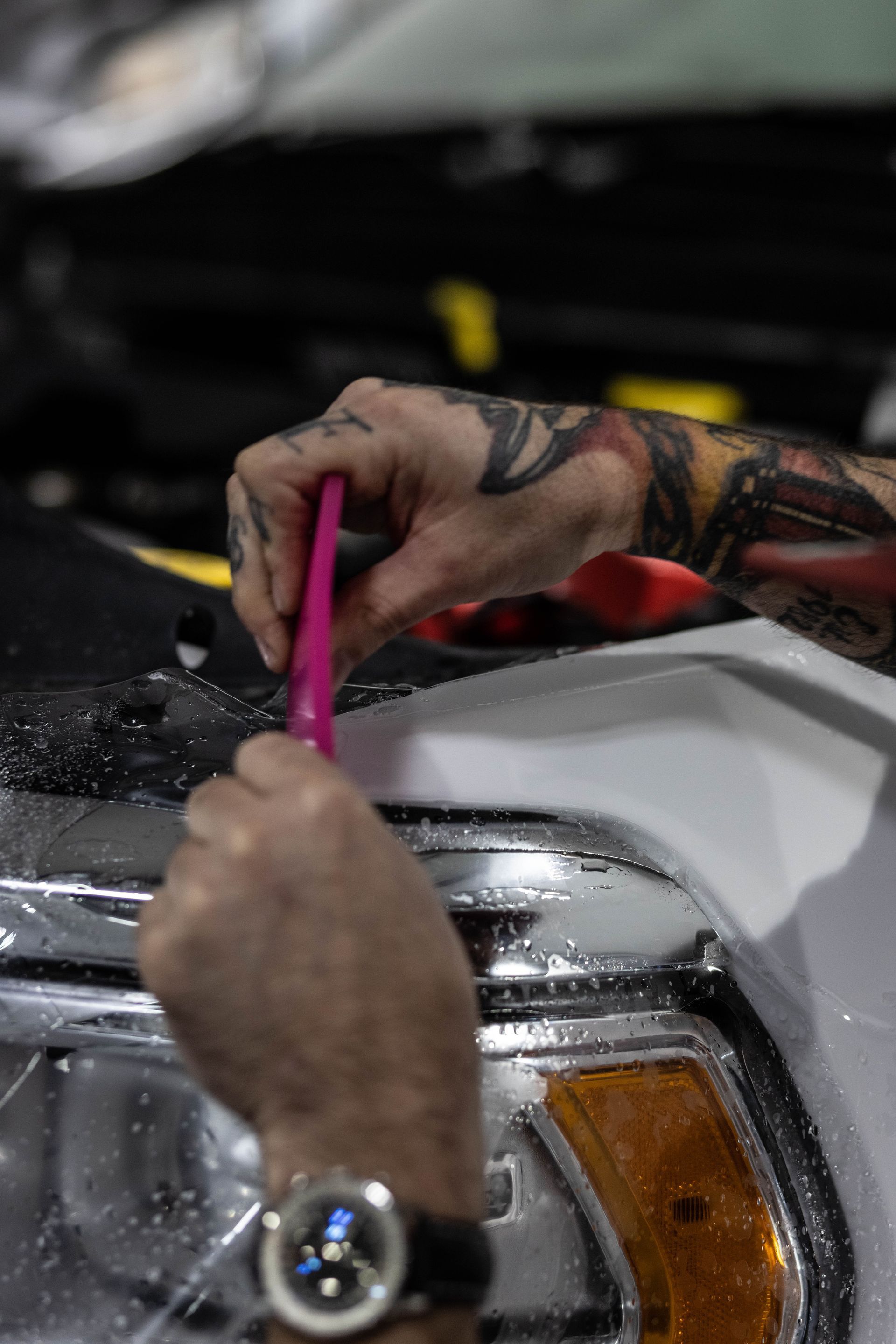 Hands using a pink brush to clean a white car headlight in a garage.