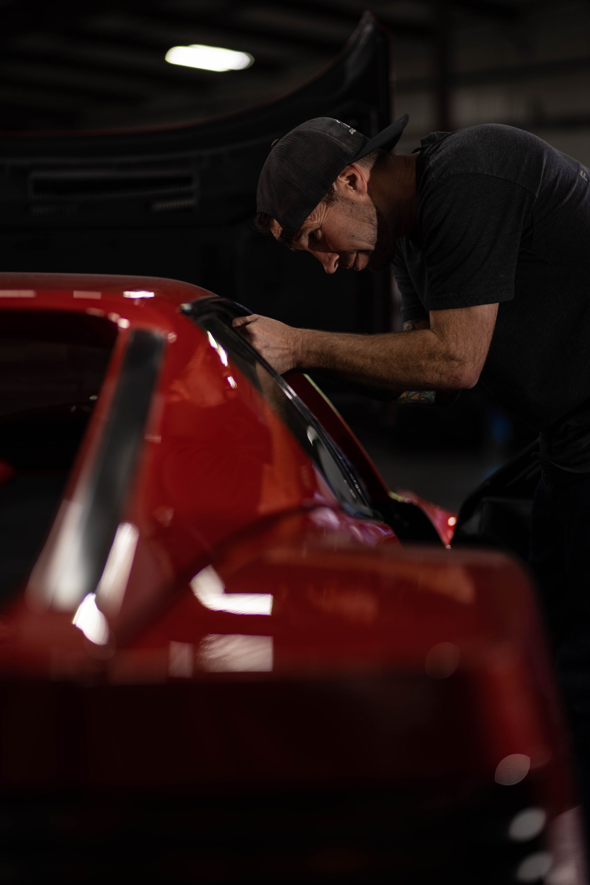 Person leaning over a red sports car in a dim garage, inspecting or working on it.