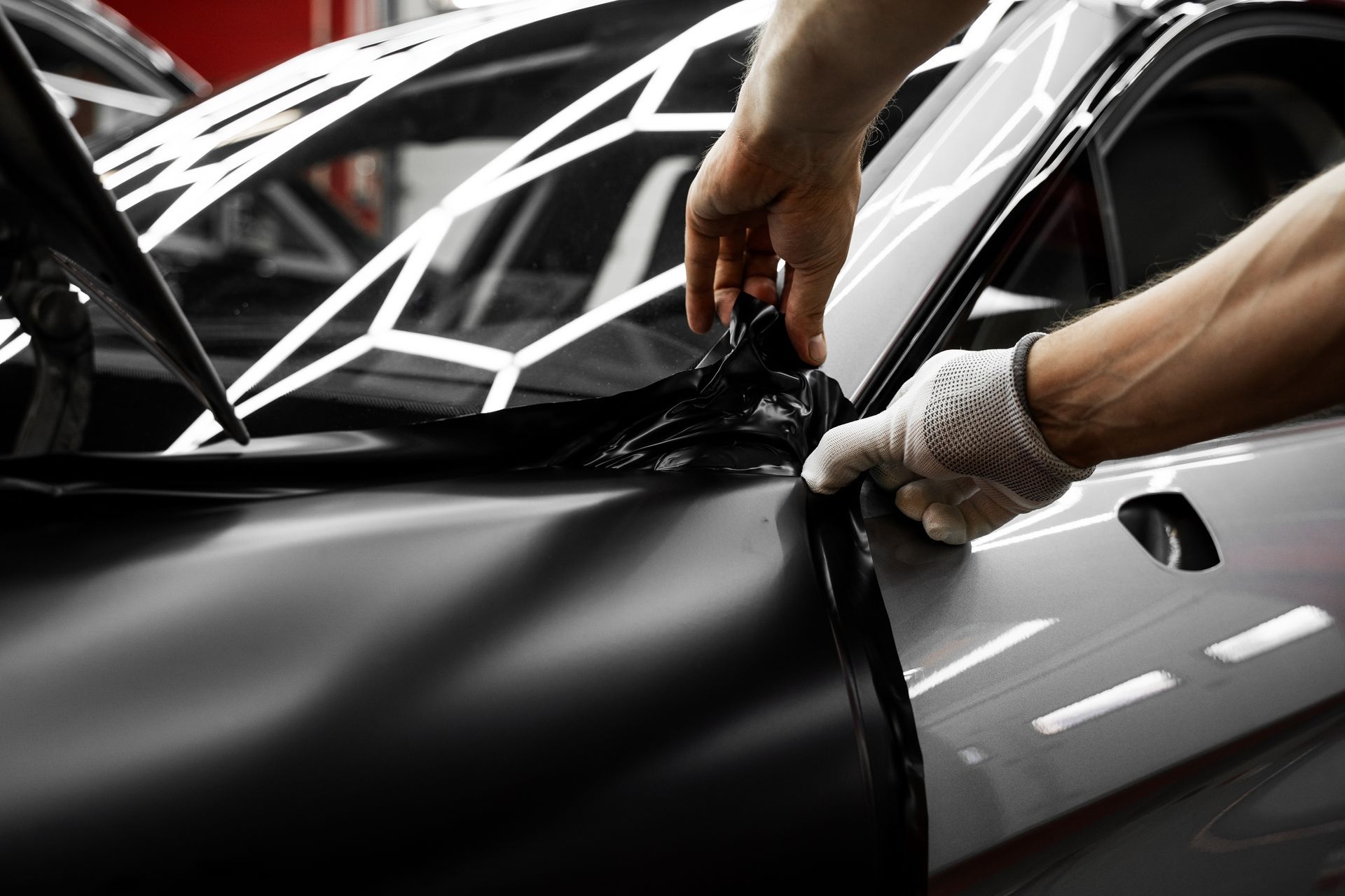 Hands applying black vinyl wrap to a silver car door in a workshop
