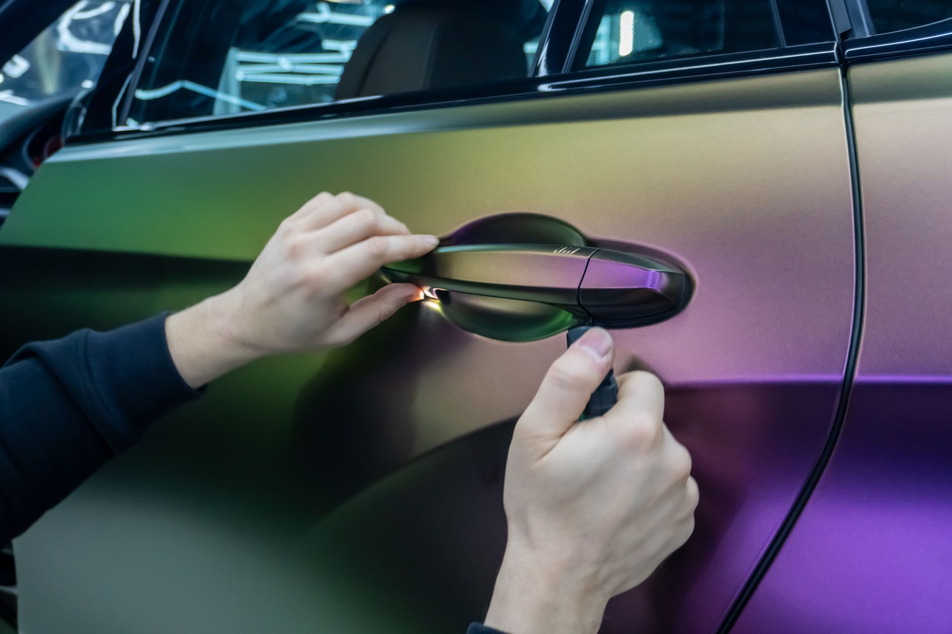 Hands applying vinyl wrap to a car door handle with green and purple reflections
