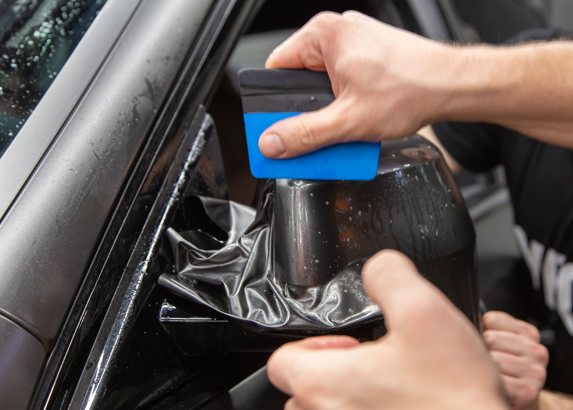Hand using a squeegee to smooth black window tint on a car window