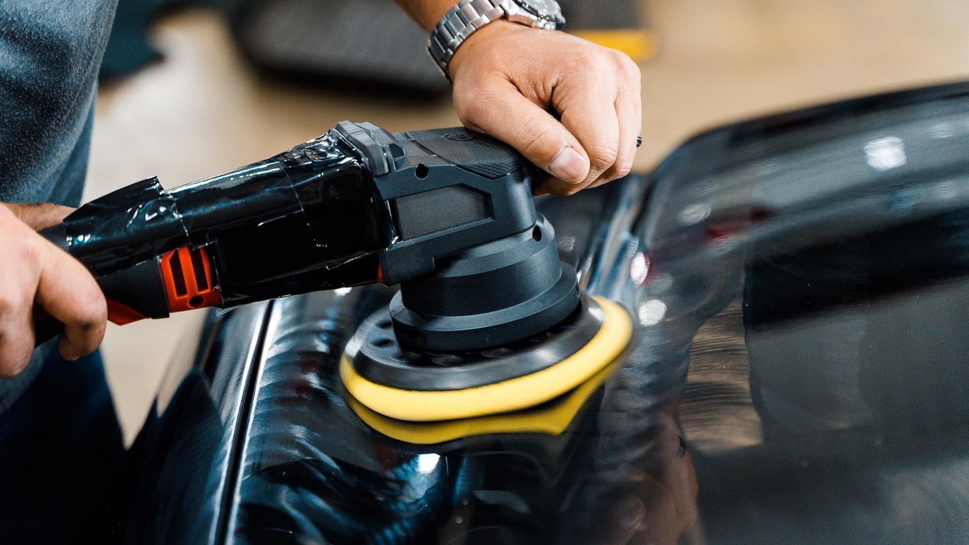 A person uses a power buffer to polish a black car’s surface.