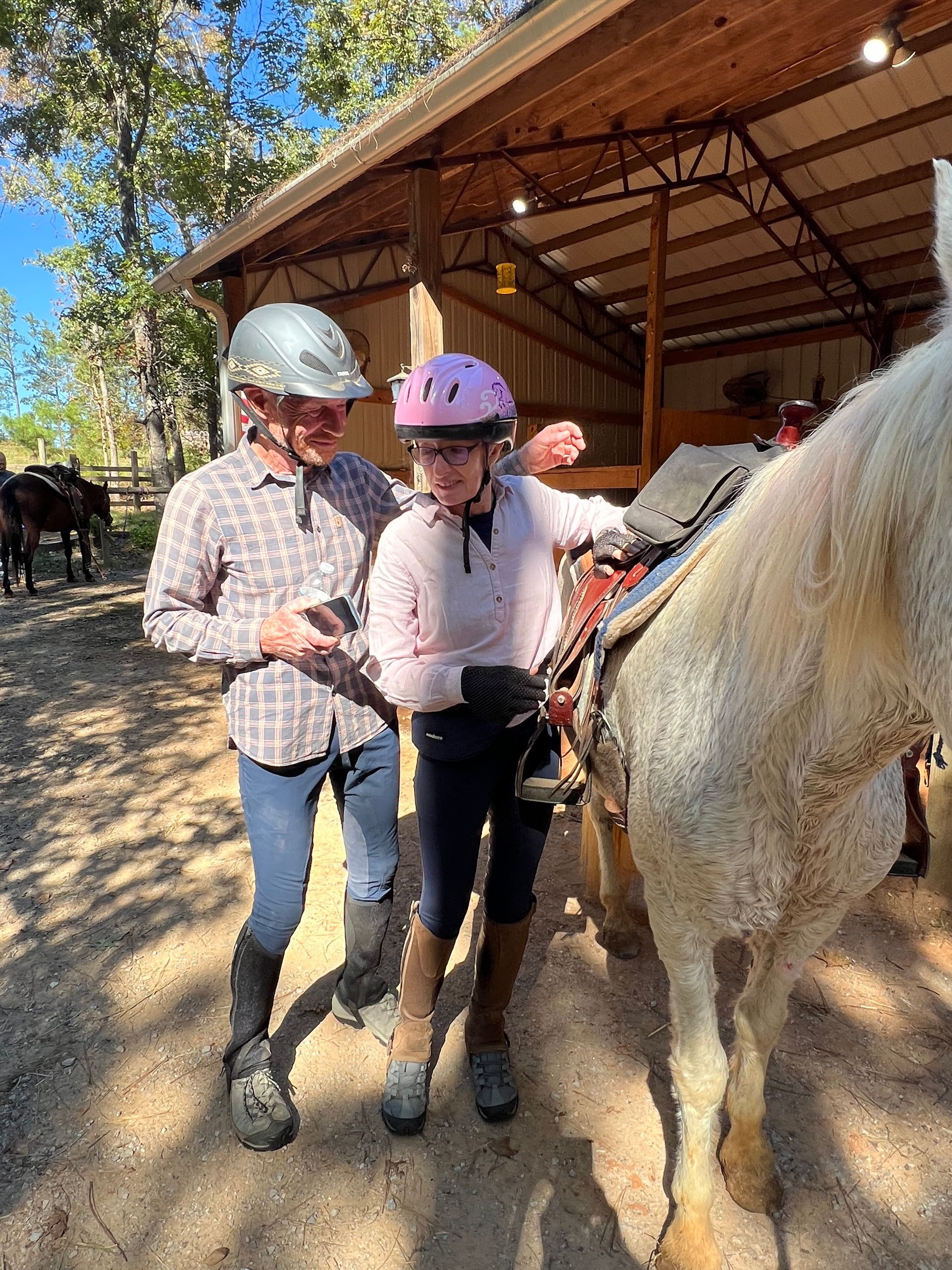Person helping another mount a horse; both wearing helmets. Outdoors, near a barn with other horses visible.