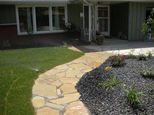 Curved stone walkway to a house entrance, bordered by green lawn and dark mulch with small plants