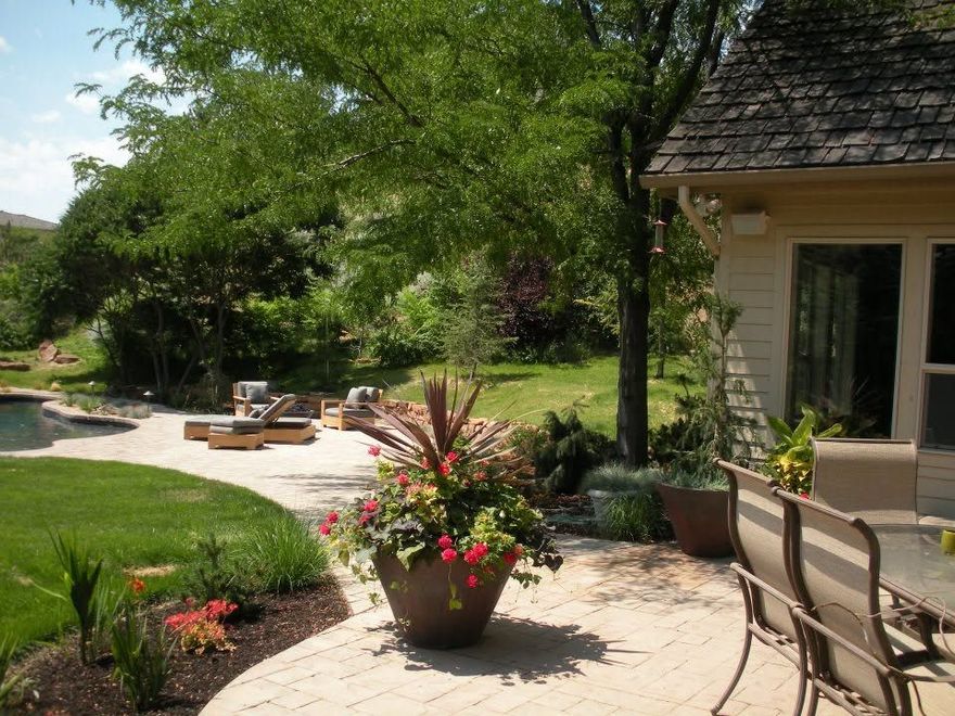 Patio with potted flowers, lounge chairs, and a house beside a green garden under trees