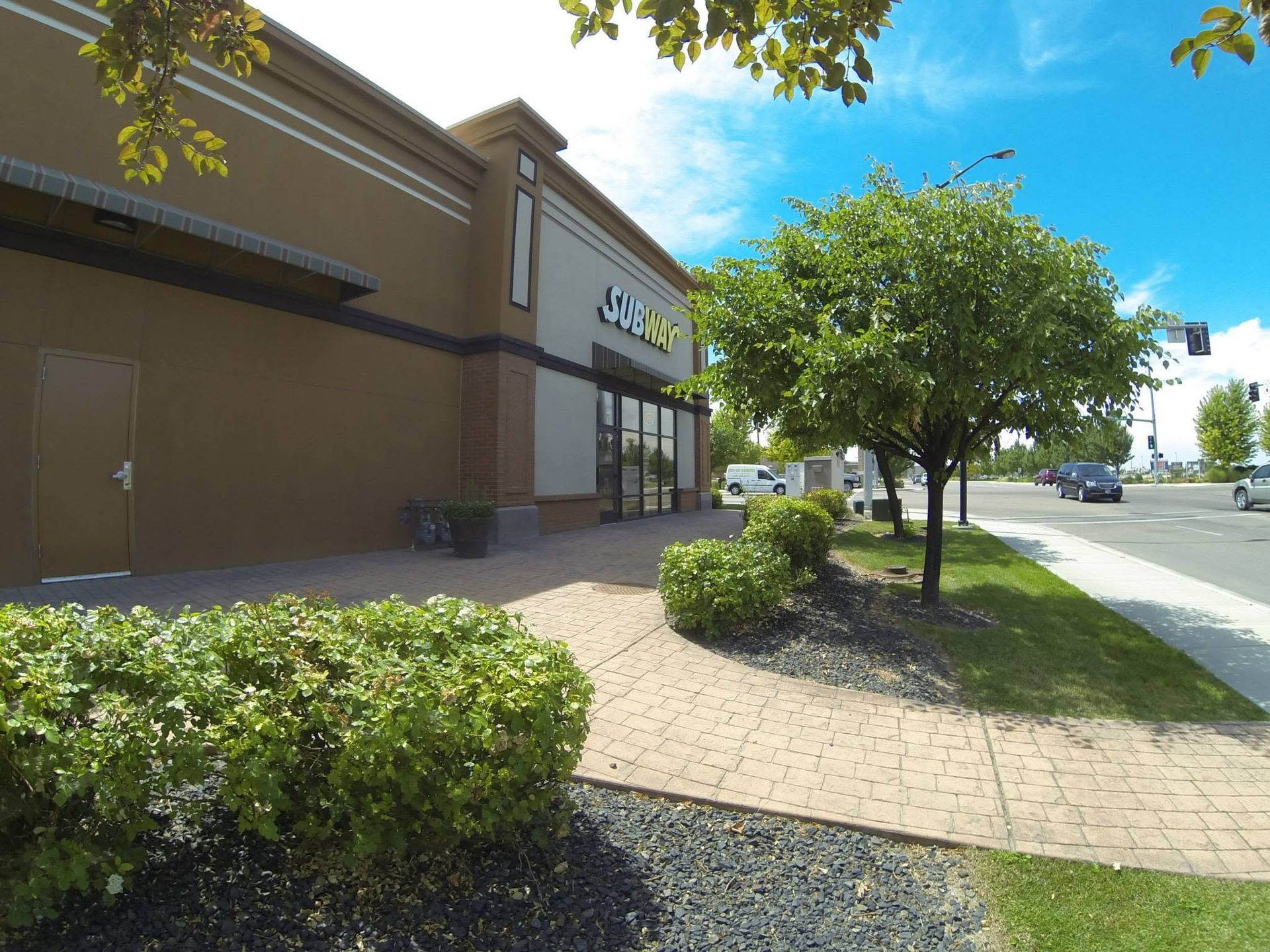 Sidewalk beside a tan storefront with shrubs, a tree, and a sunny street