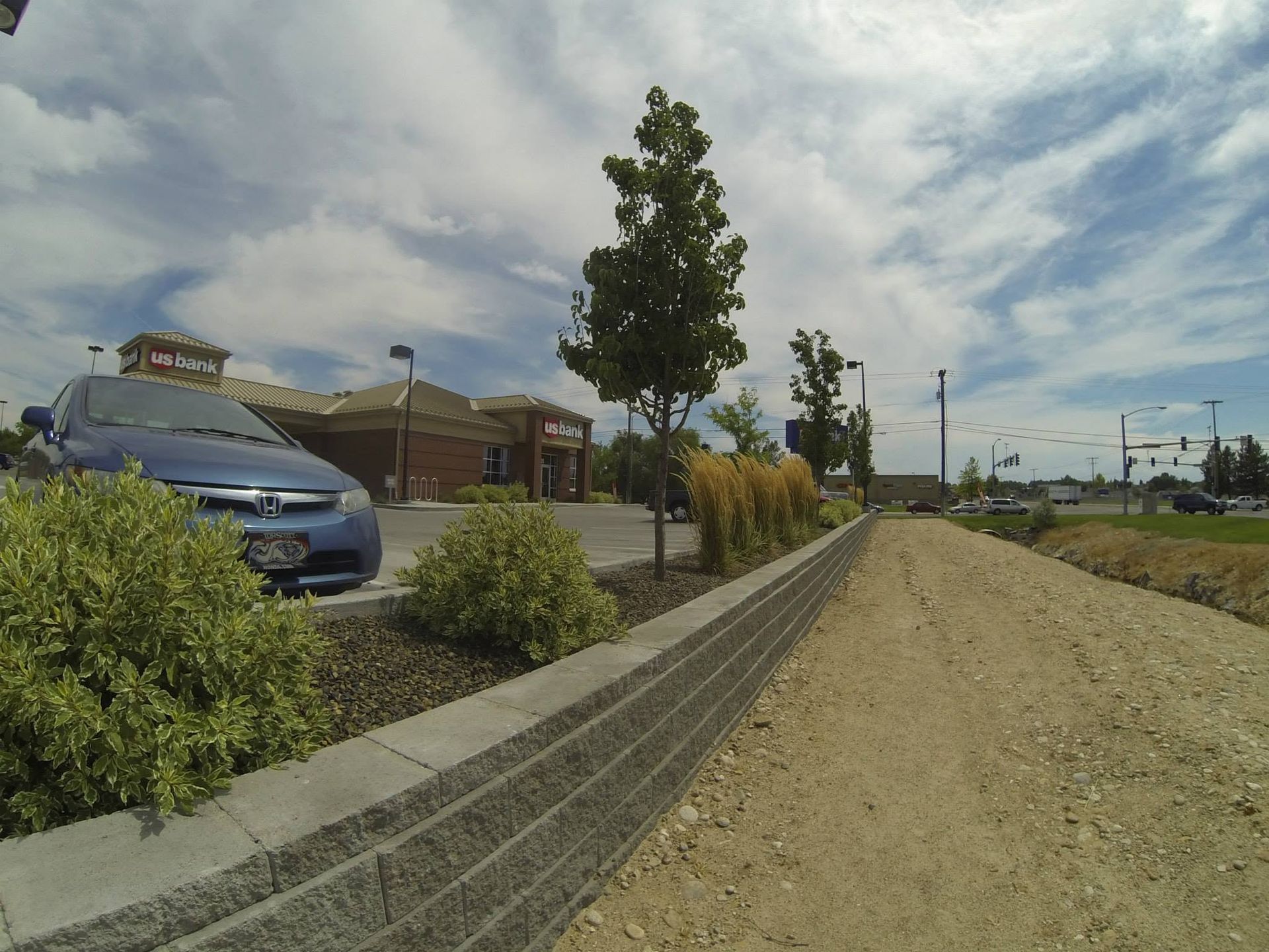 Gravel path beside parked cars and brick buildings under a cloudy sky, with a tree and sidewalk barrier