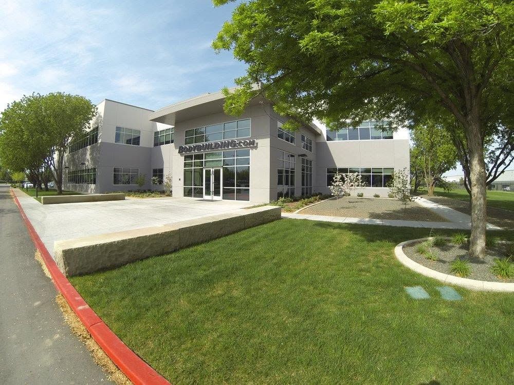 Modern white building with glass entrance, trees, and a landscaped lawn beside a curb