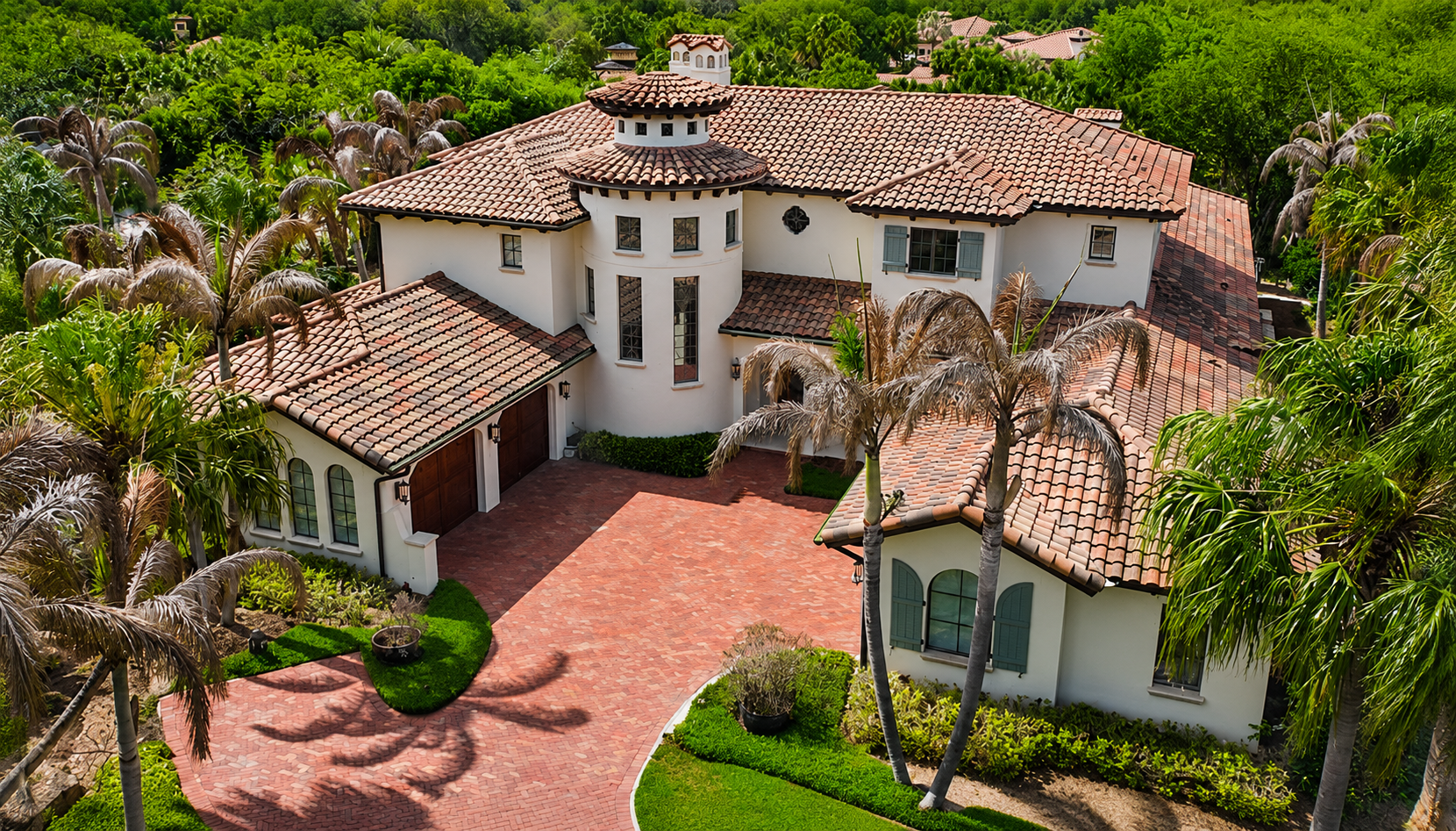 Aerial view of a large Mediterranean-style villa with red tile roofs, palm trees, and a brick driveway