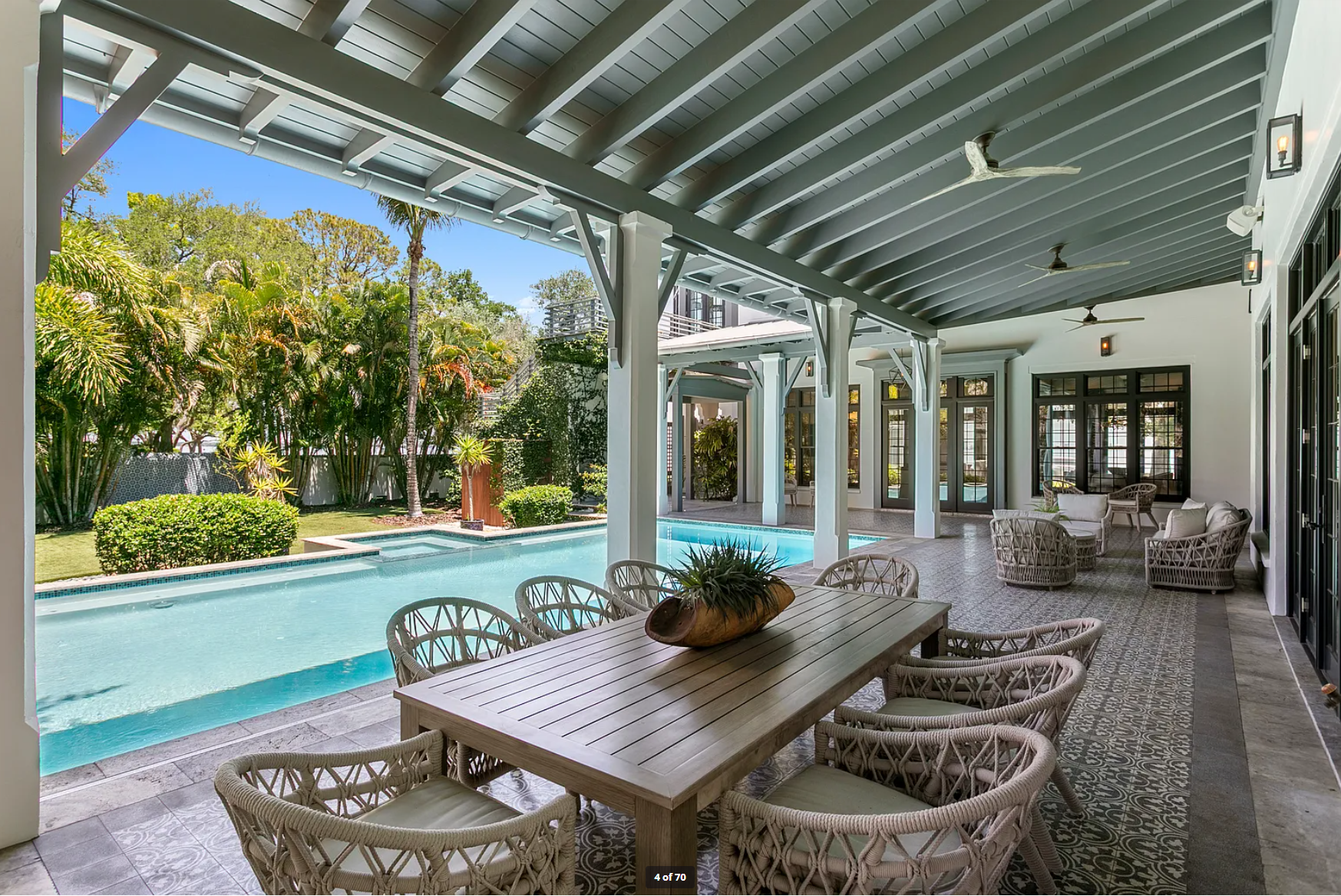 Covered patio with wicker dining table beside a turquoise pool and garden view