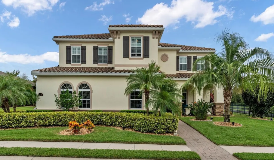 Two-story beige house with a tile roof, palm trees, and a manicured lawn under a blue sky