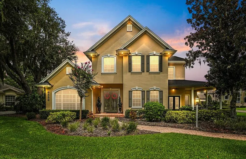 Two-story suburban house with warm exterior lights at dusk, framed by trees and a landscaped lawn