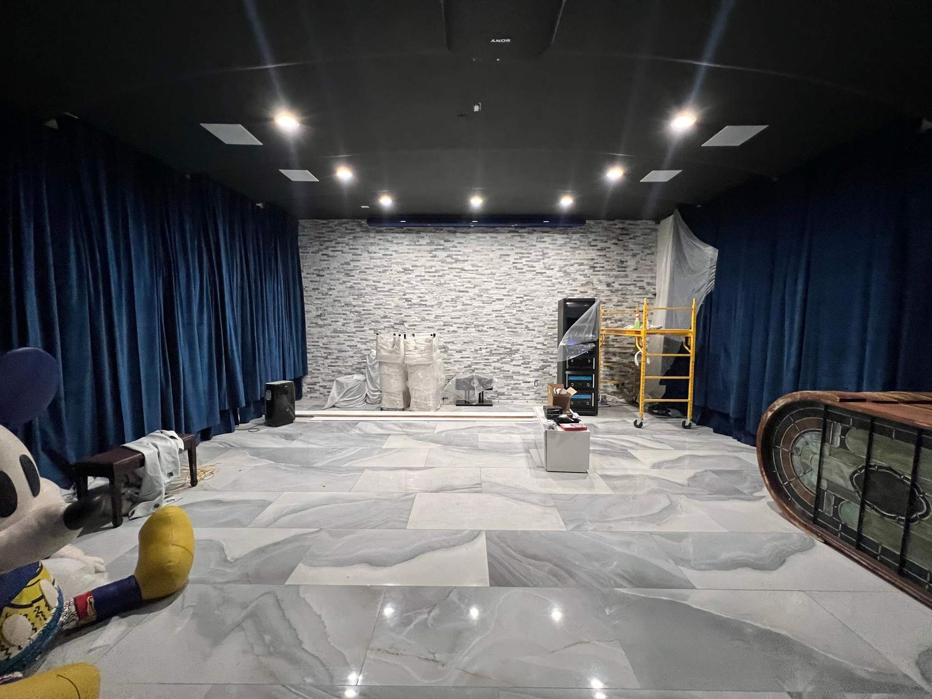 Empty room with stone wall, dark curtains, overhead lights, and scattered props on a tiled floor