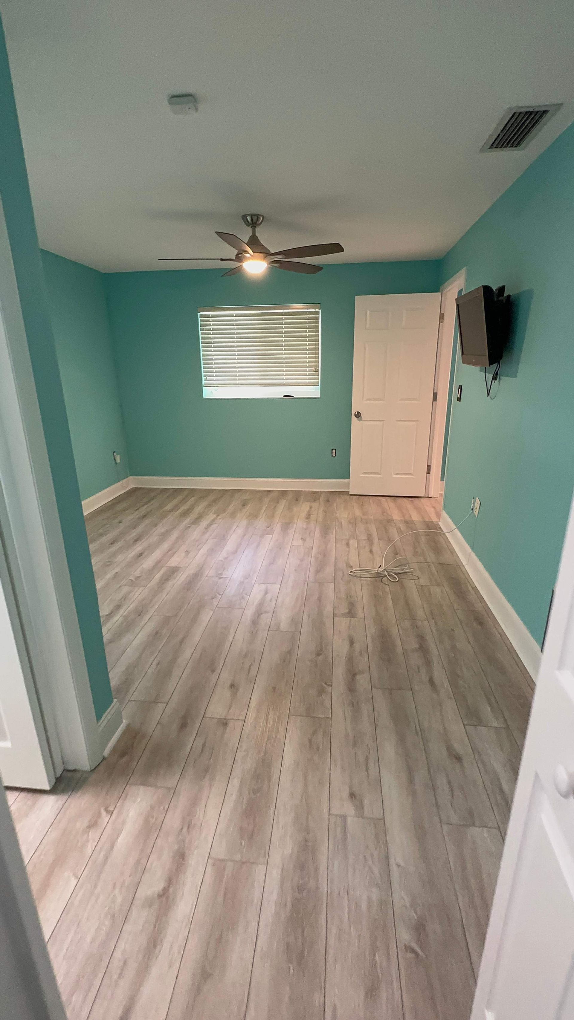 Empty turquoise bedroom with wood floors, ceiling fan, window blinds, and a closed white door.