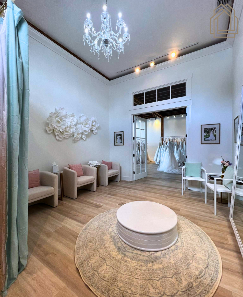 Elegant white boutique room with chandelier, pale wood floor, round ottoman, and bridal gowns by the doorway
