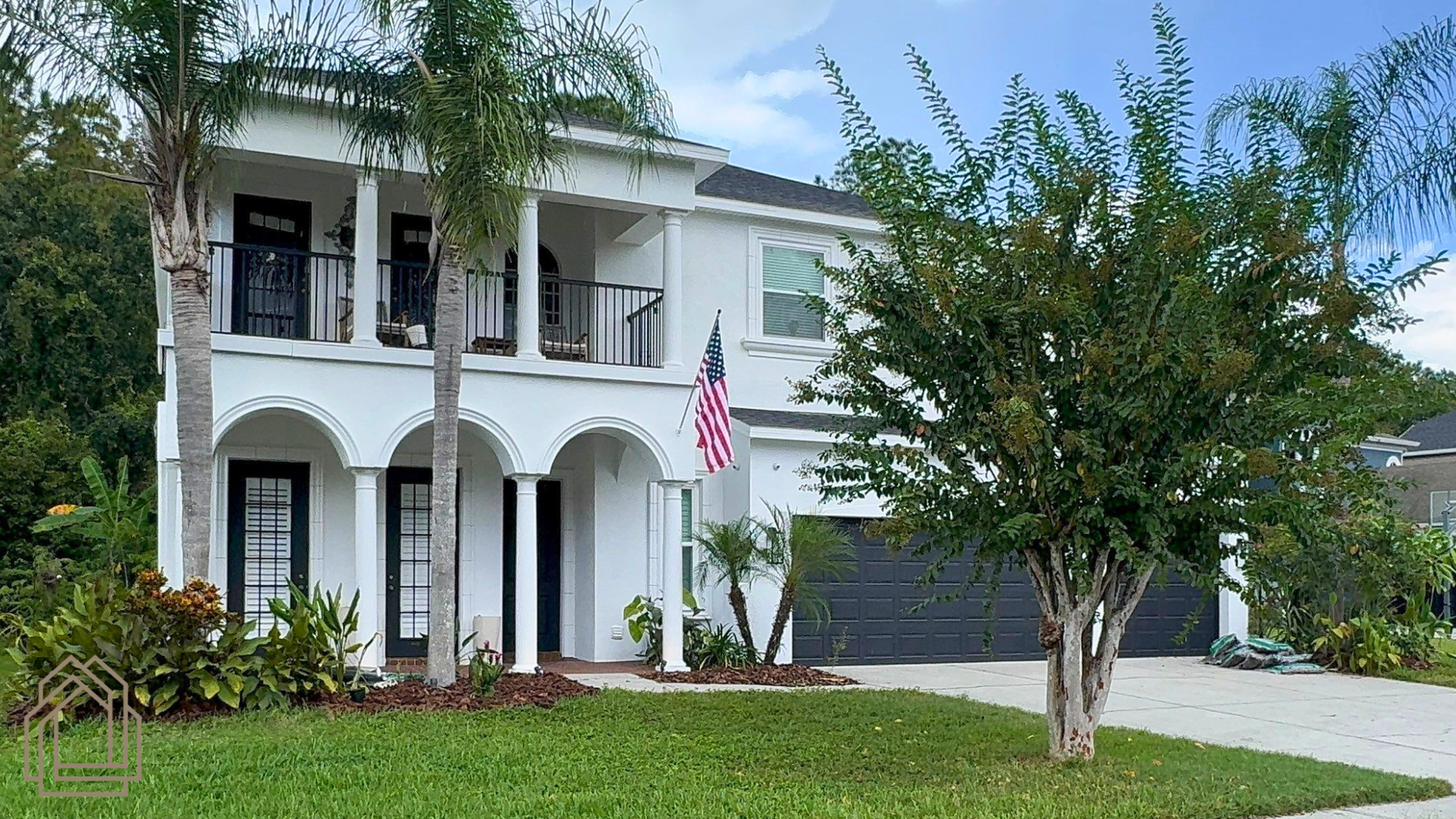 Two-story white house with arched porch, palm trees, and an American flag by a green lawn