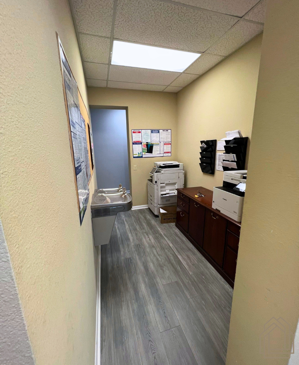 Narrow office hallway with beige walls, gray floor, copier, cabinets, and bulletin board under fluorescent lights