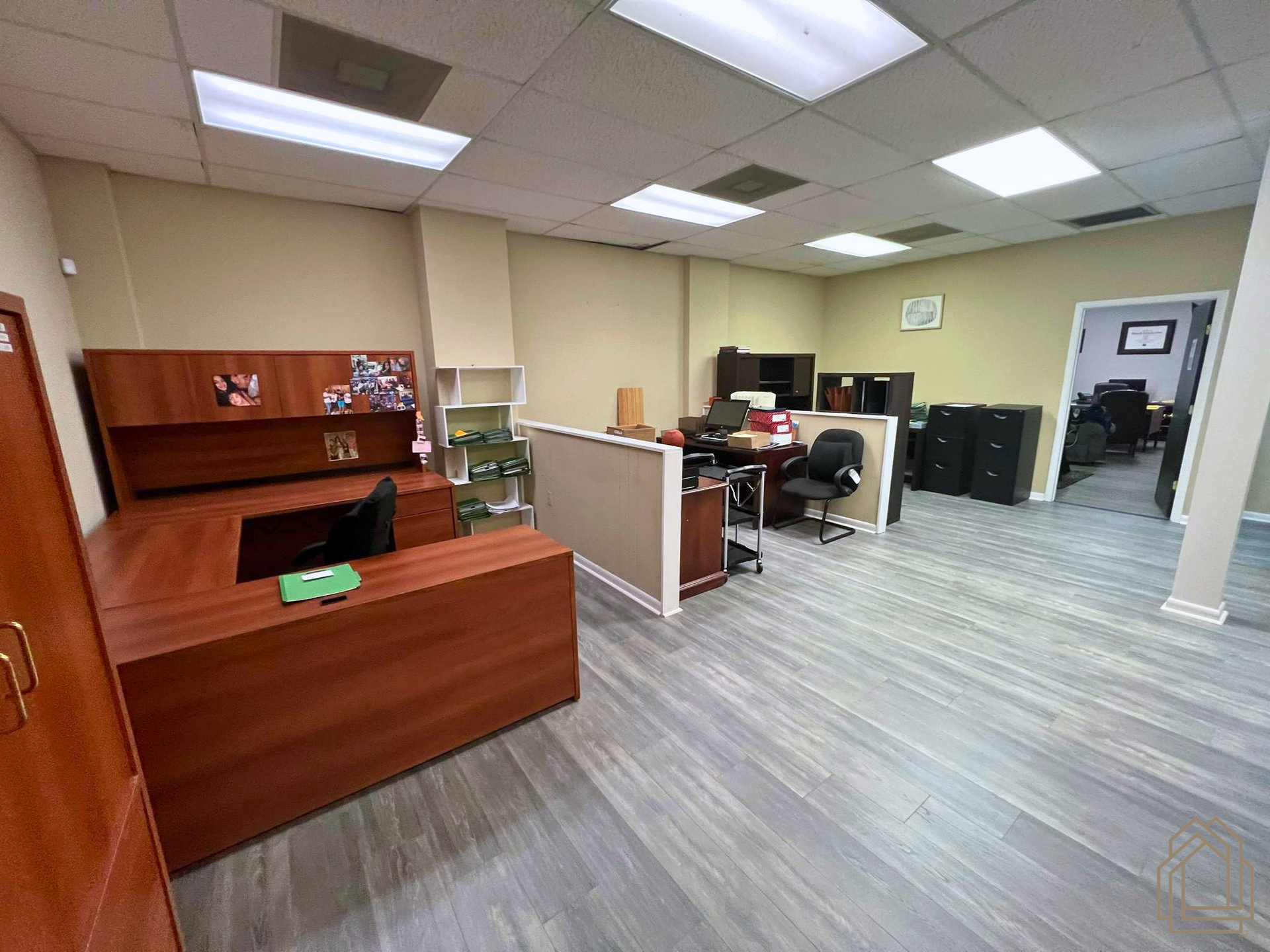 Office reception area with wooden desk, cubicle workspace, and gray carpet under fluorescent lights