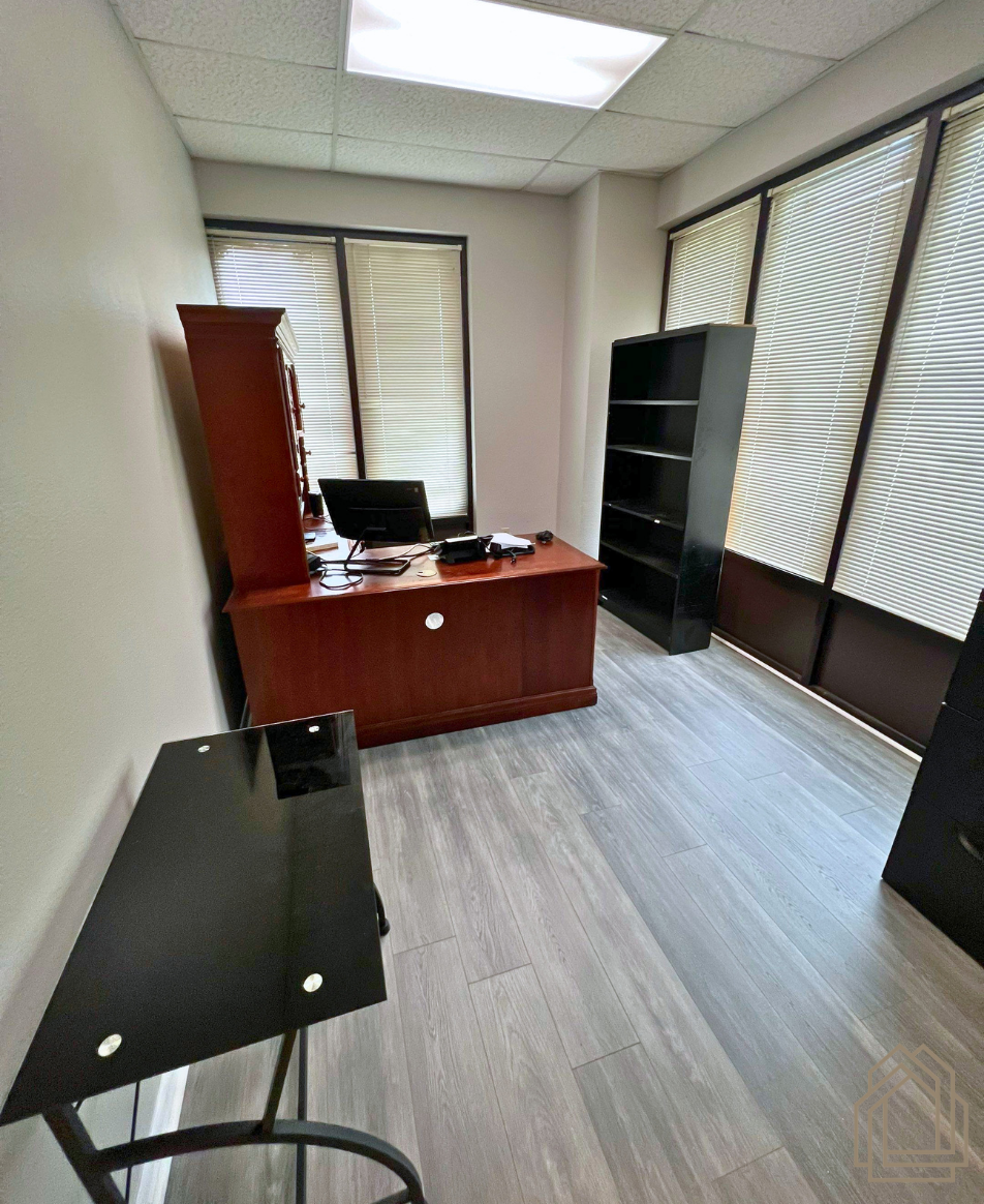 Small office with a wooden desk, computer, black shelving, and large windows with blinds.