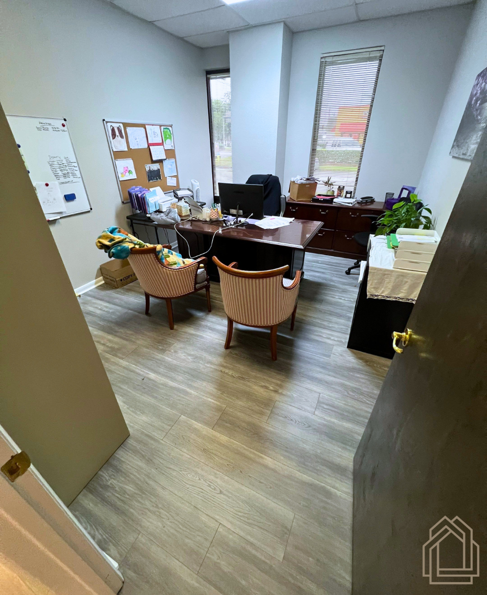 Office with desk, computer, chairs, bulletin board, and window blinds; plants and papers on the desk.