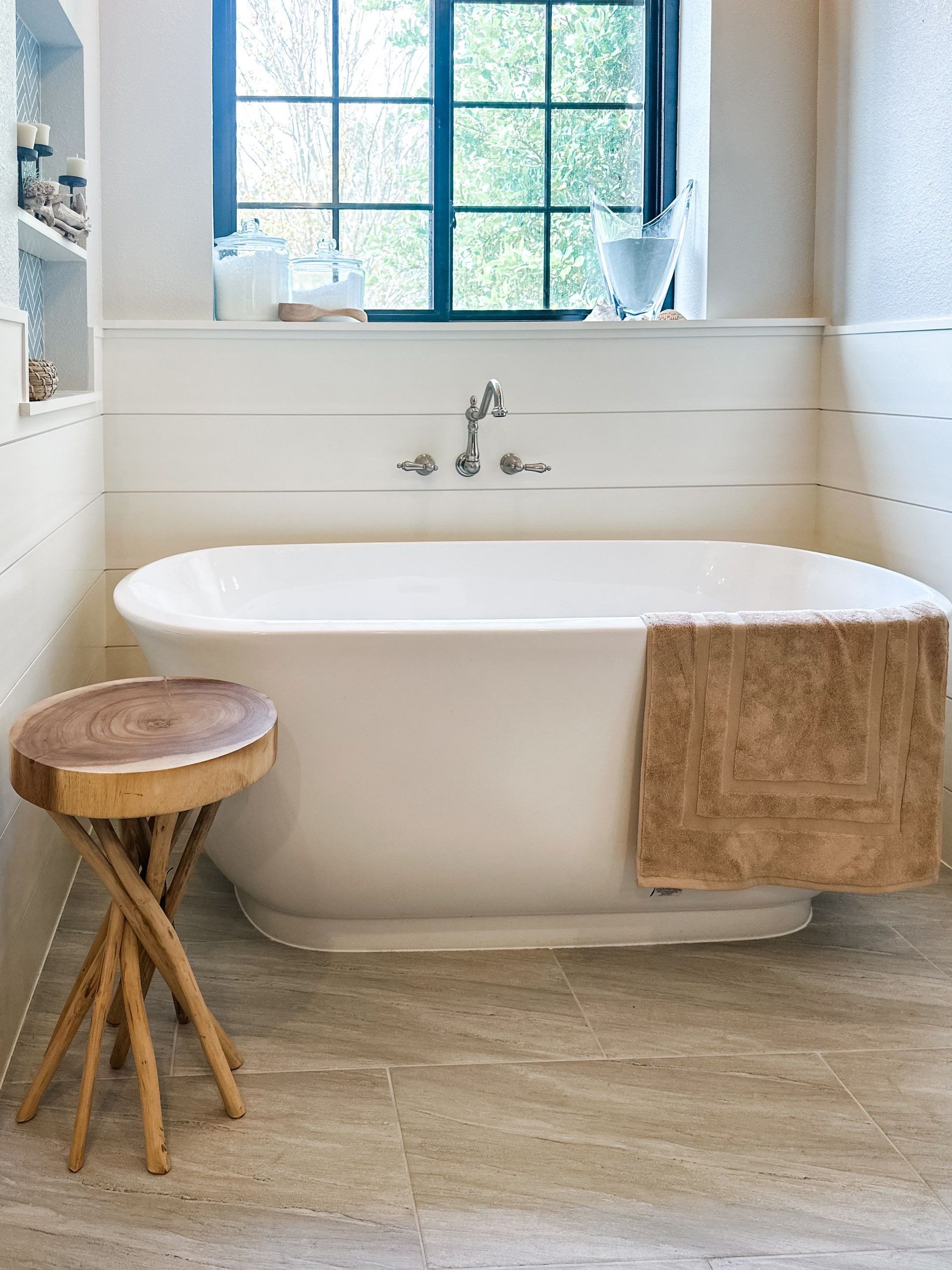 Bright bathroom with a white freestanding tub, wooden stool, tan towel, and window above the sink.
