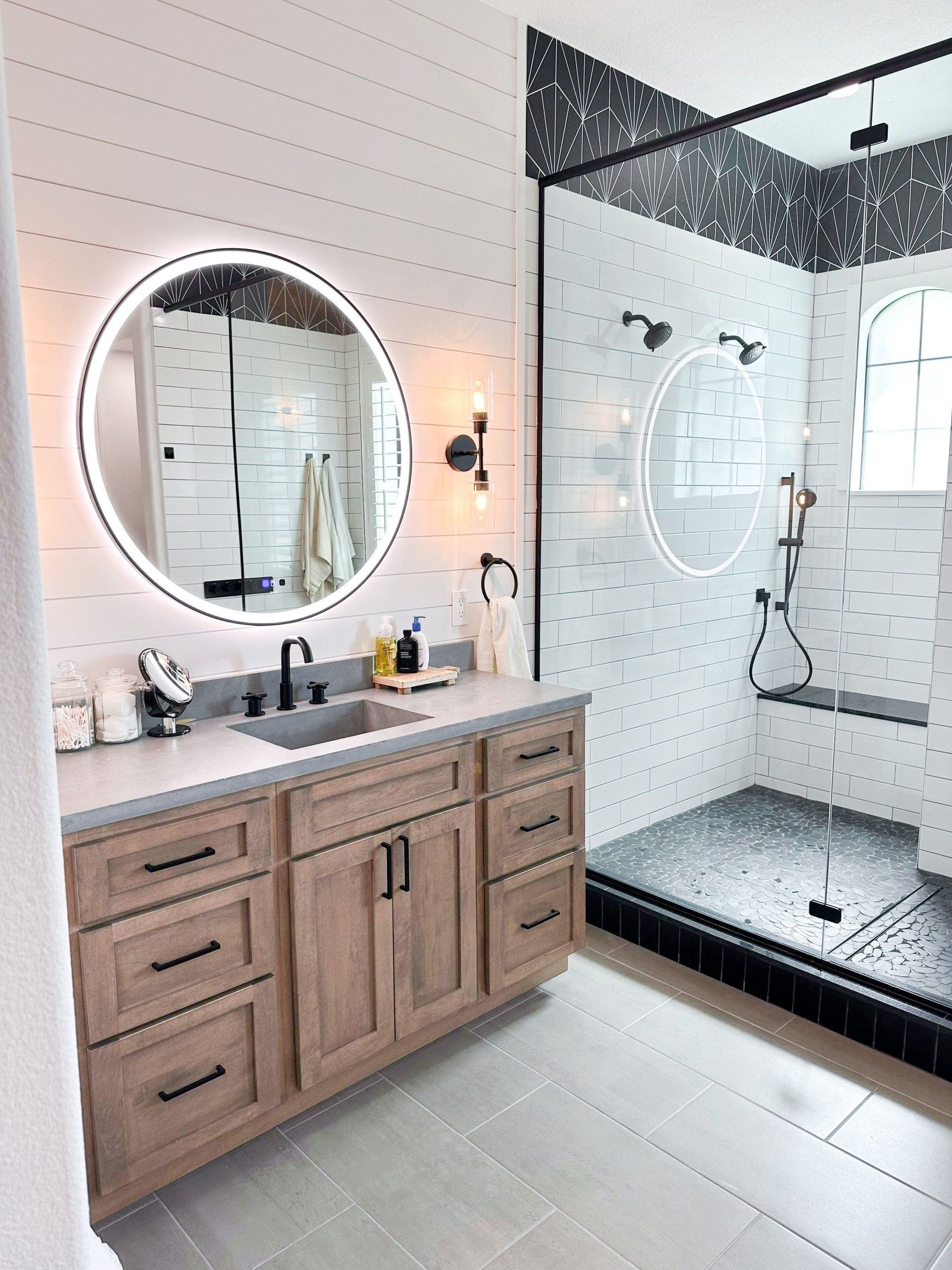 Modern bathroom with wood vanity, round mirror, white tile, and black-framed glass shower.