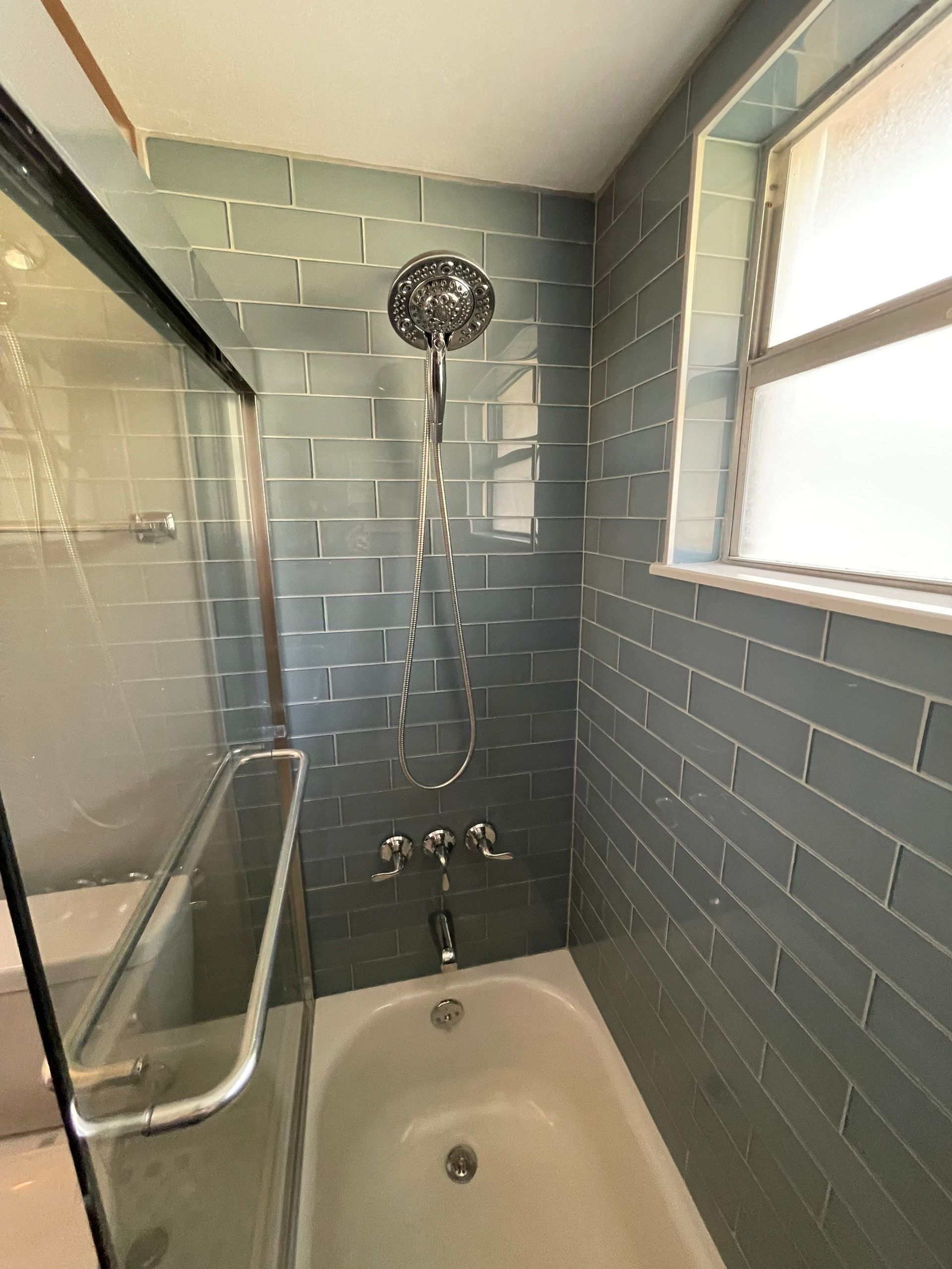 Small bathroom with blue tile, a bathtub, wall-mounted showerhead, grab bars, and a window.