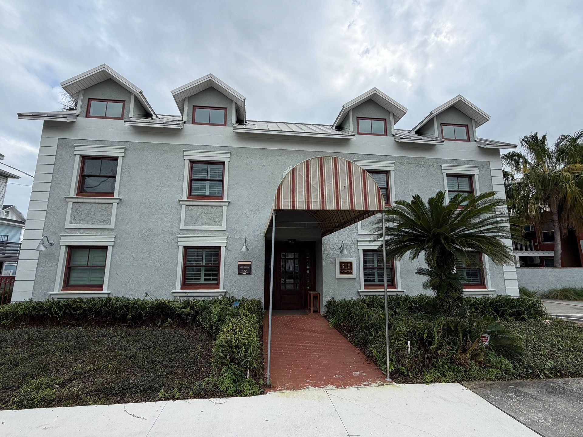 Front view of a pale green building with striped awning, brick path, and palm trees