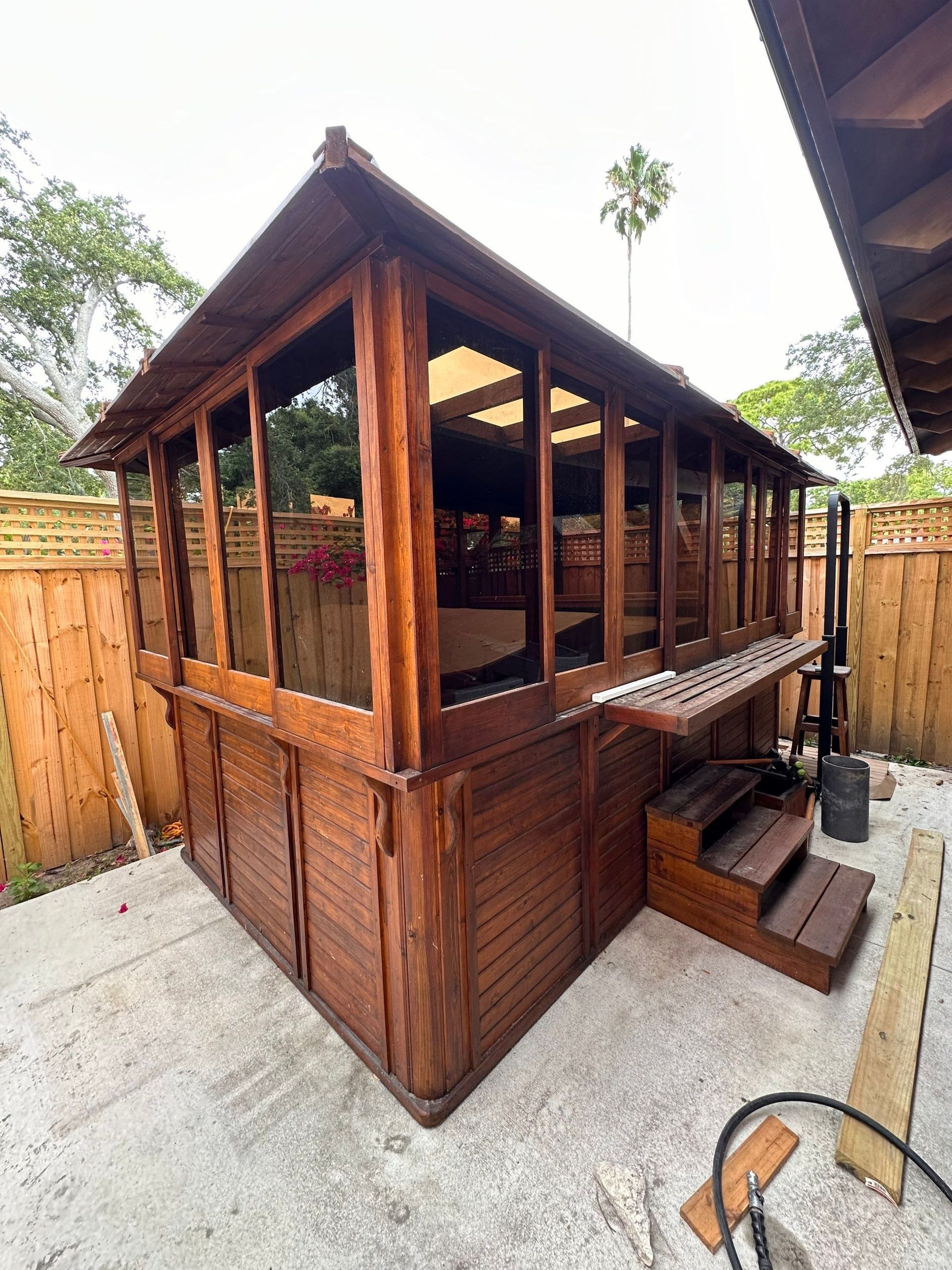 Wooden outdoor gazebo with open windows and steps in a fenced backyard courtyard.