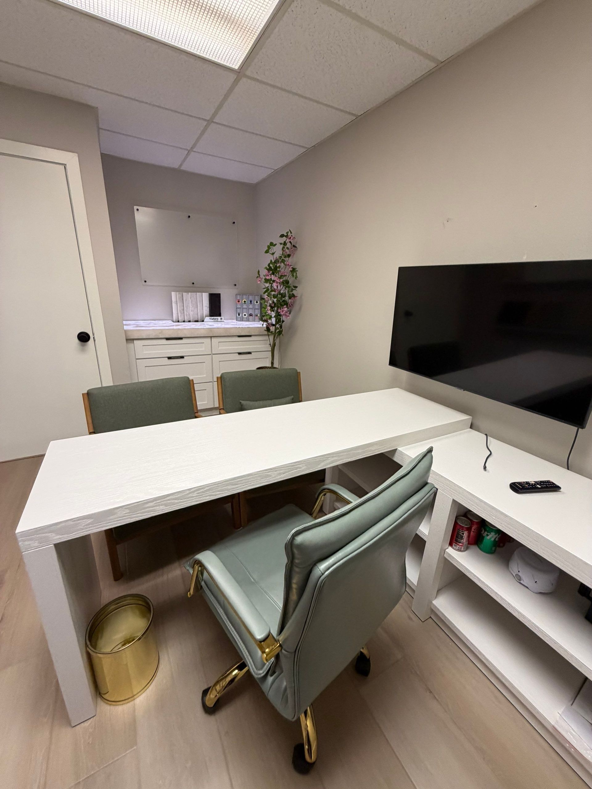 Small modern office with white desk, teal chair, wall-mounted TV, and windowed kitchenette nook.