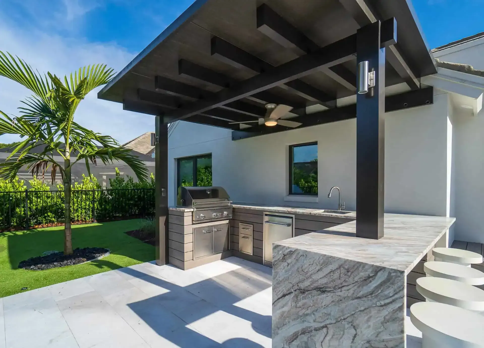 Outdoor patio kitchen with built-in grill, marble counter, and pergola roof in a sunny backyard