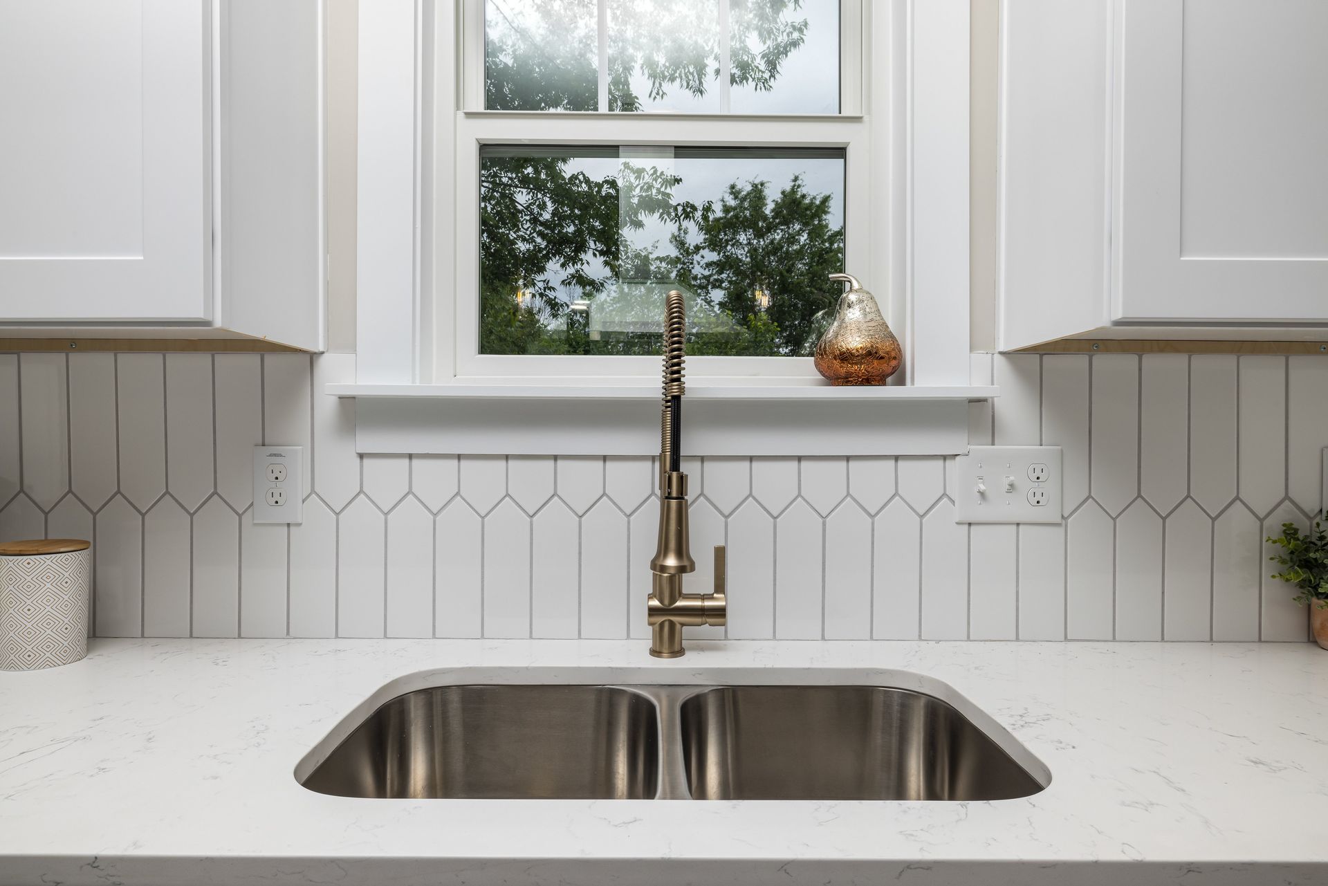 Modern kitchen sink beneath a window, with white cabinets, brass faucet, and light countertop.
