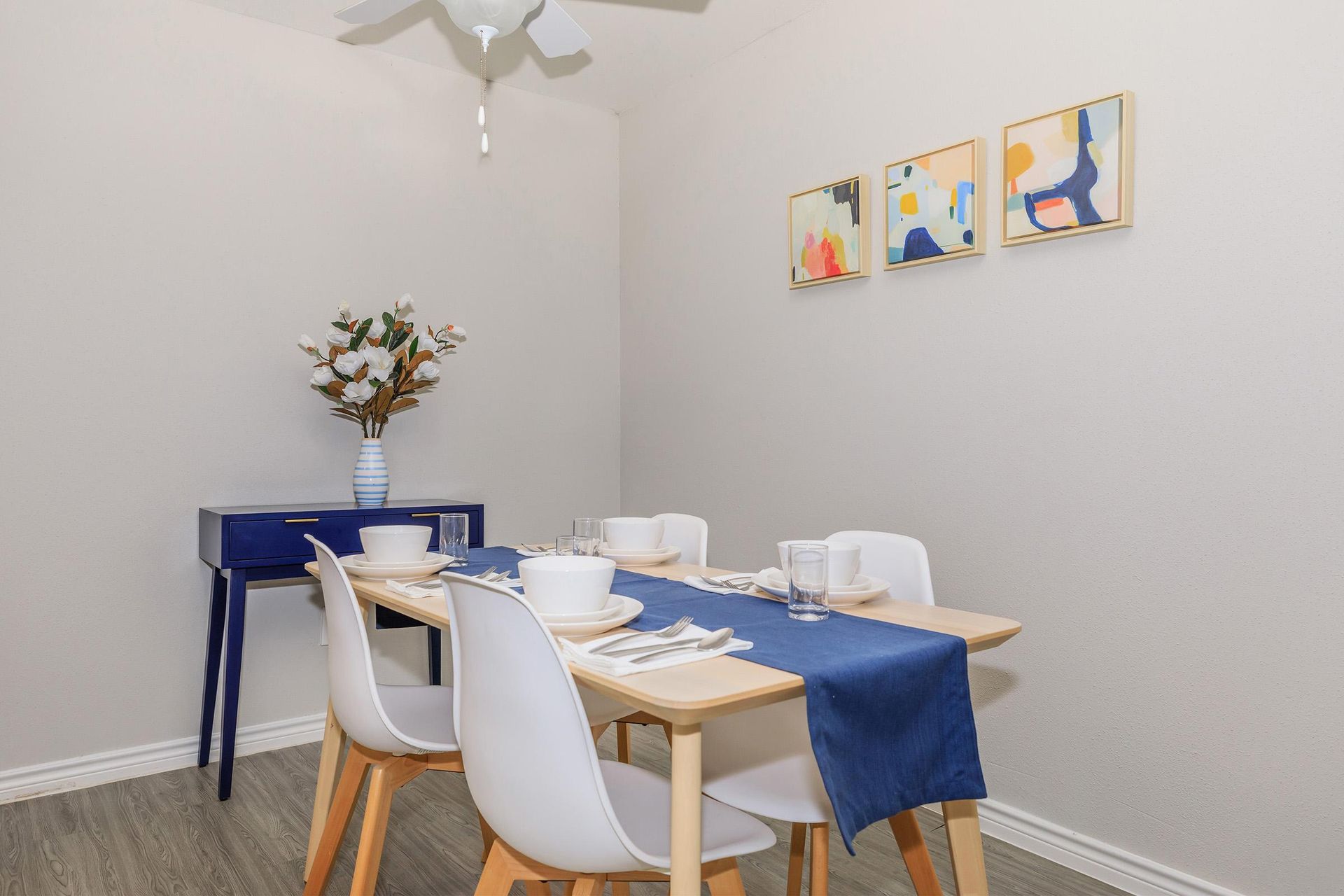 Dining area in an apartment with a light wood table, white chairs, a blue runner, and abstract wall art.