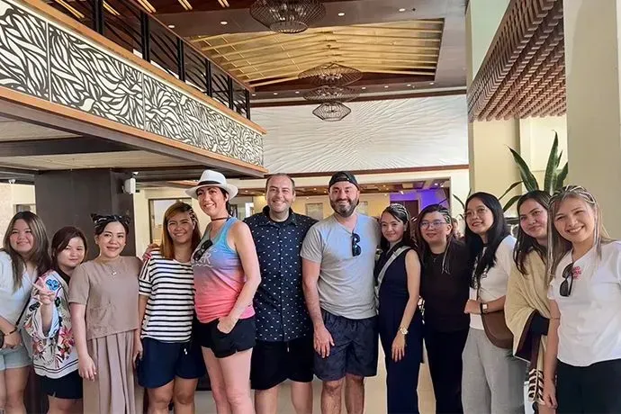Group of smiling people posing indoors in a bright lobby with wood ceiling and decorative balcony.