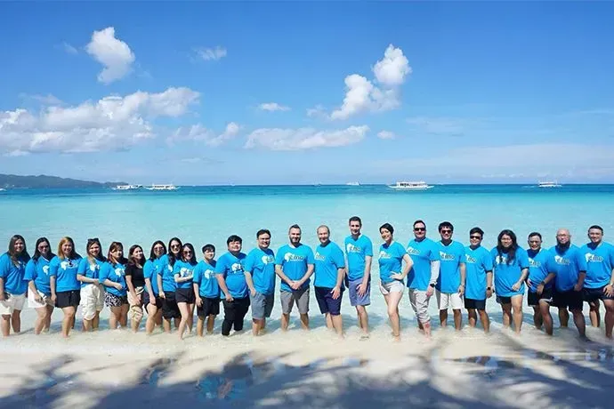 Group of people in blue shirts standing on a sunny beach with ocean and sky behind them