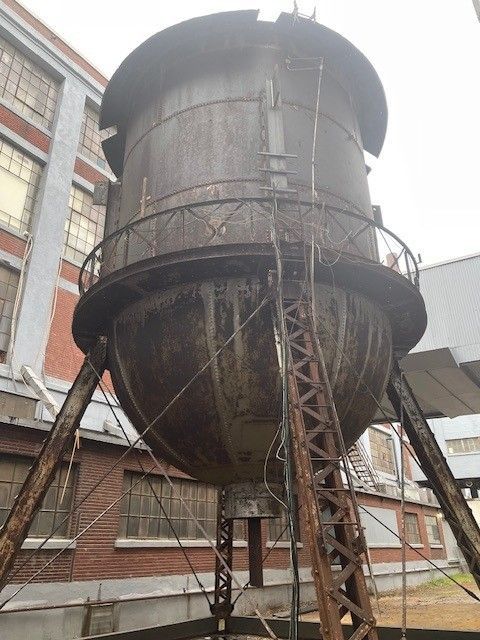 Rusty industrial water tower on stilts beside a brick building.