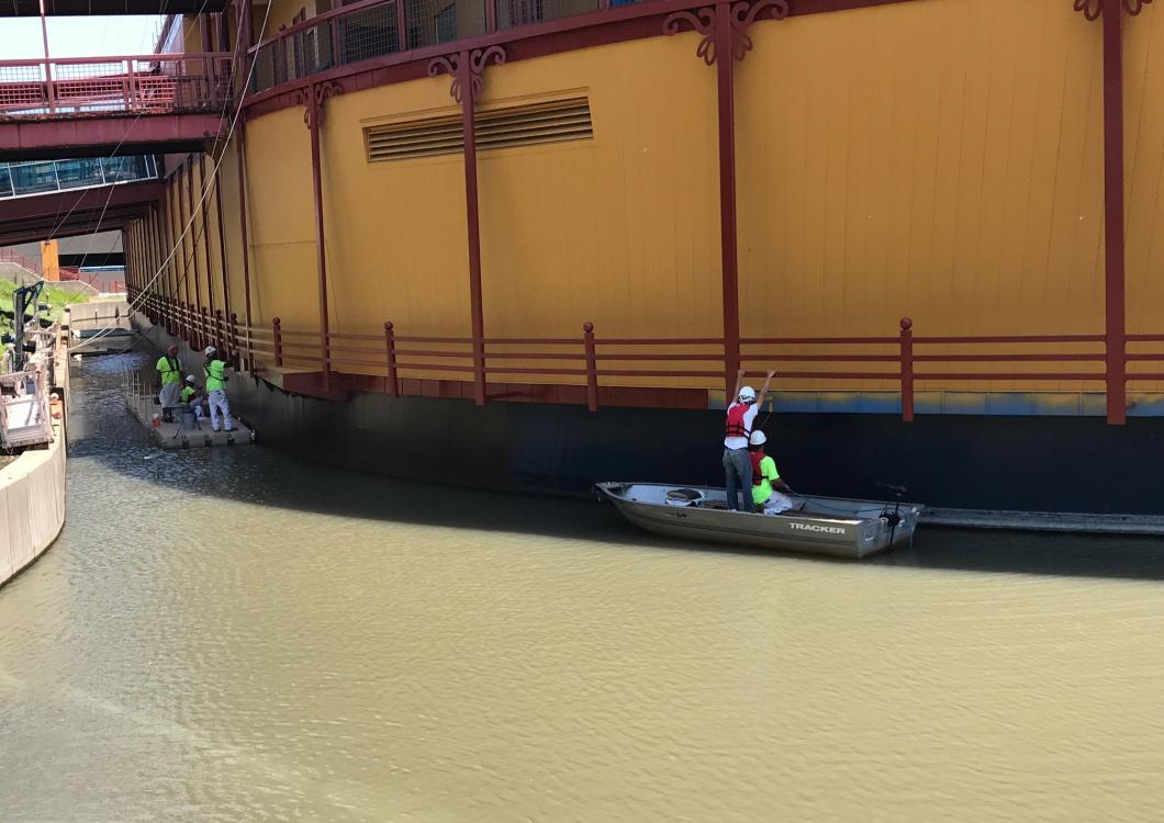 Two people in a small boat on a canal beside a large orange bridge or wall in daylight