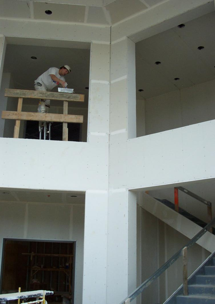Worker on a scaffold inside a unfinished building, painting the upper wall near a staircase.